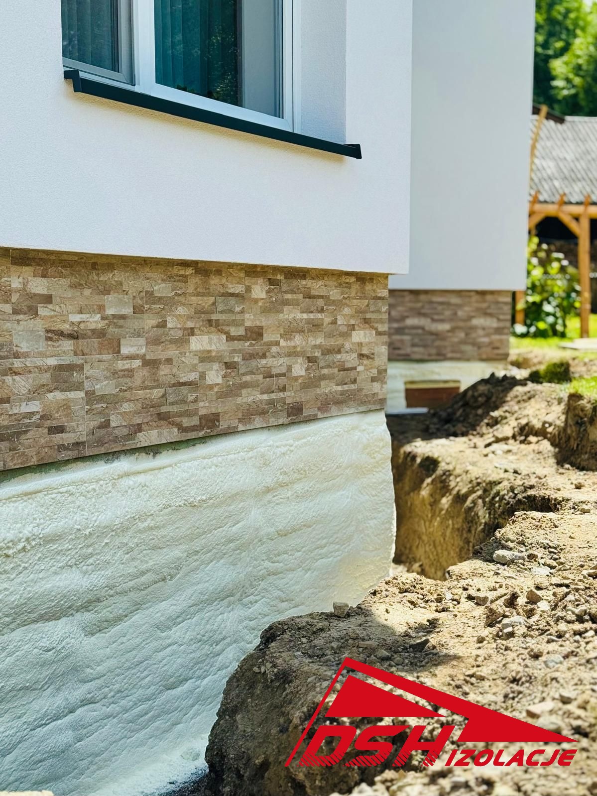 Ocieplenie fundamentu pianką PUR, widoczne wykopane fundamenty, nałożona pianka, fragment elewacji z oknem i dekoracyjną okładziną.
