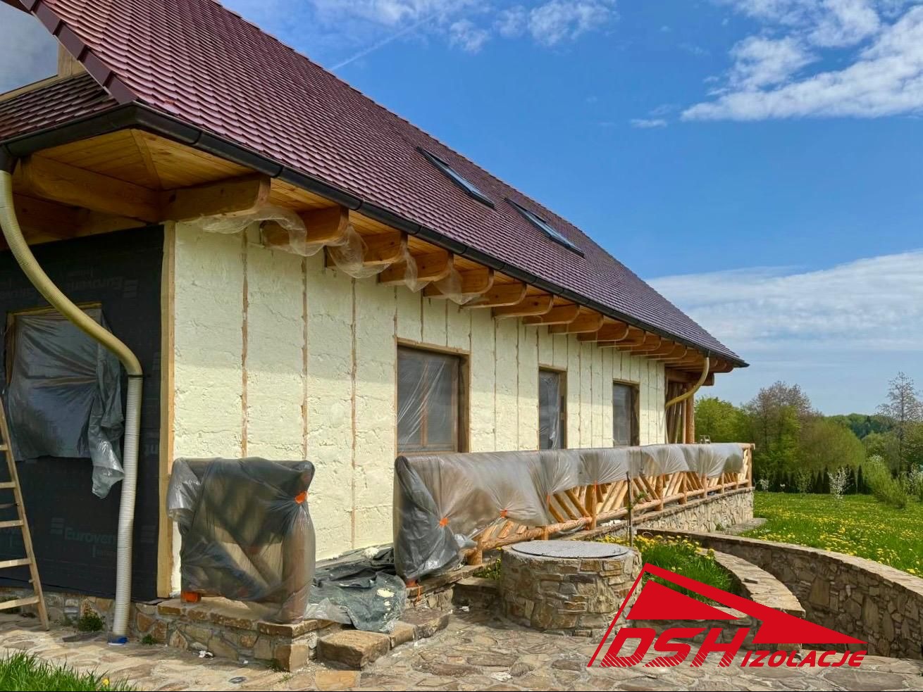 Dom jednorodzinny w trakcie ocieplania ścian zewnętrznych pianką PUR, widoczne zabezpieczone okna folią i fragment elewacji z nałożoną pianą. Słoneczny dzień.