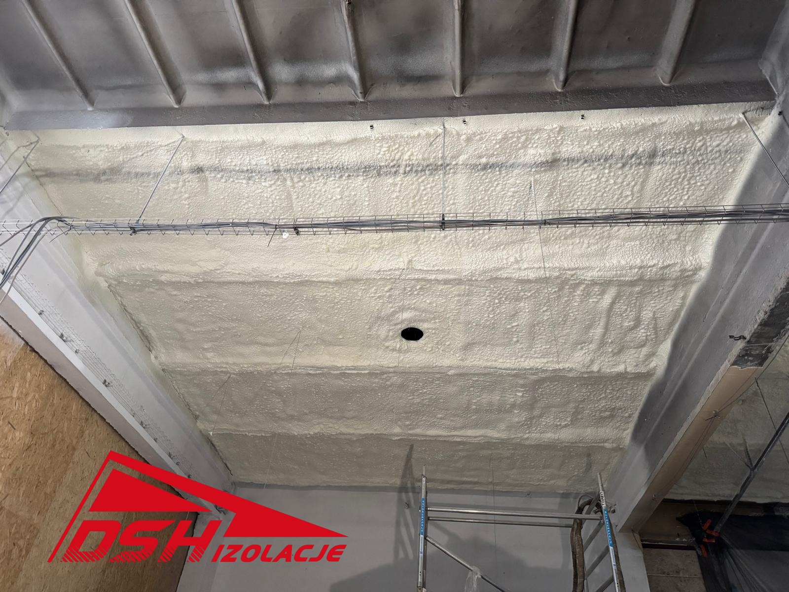 Poddasze po ociepleniu pianką PUR, widoczne równomierne pokrycie izolacją, szare belki konstrukcyjne i przewody elektryczne pod sufitem. Na dole logo firmy.
