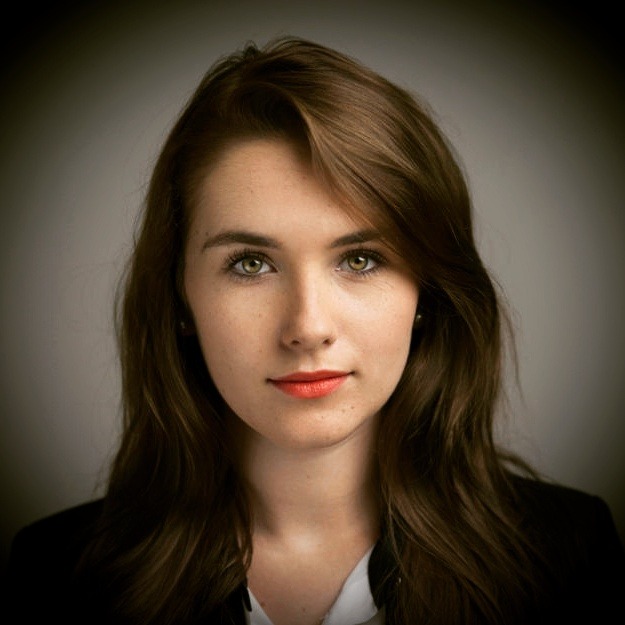 Portret młodej kobiety z brązowymi włosami i zielonymi oczami, ubrana w czarną marynarkę i białą koszulę, z delikatnym makijażem i pomadką w odcieniu koralowym, na rozmytym szarym tle.