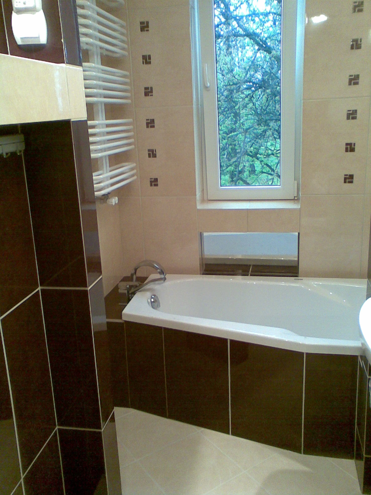 Wykończona łazienka z narożną wanną obudowaną ciemnobrązowymi płytkami, beżowe płytki na ścianach z dekoracyjnym wzorem, grzejnik drabinkowy i okno z widokiem na drzewa.