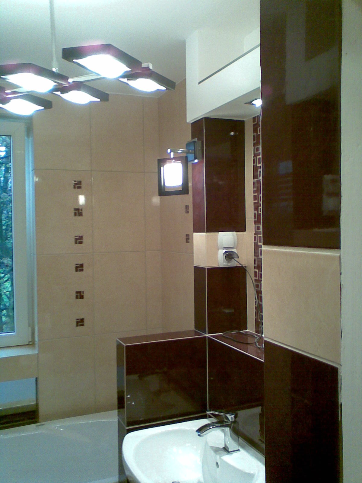 Wykończona łazienka z beżowo-brązowymi płytkami, umywalką i nietypowym oświetleniem sufitowym oraz ściennym.