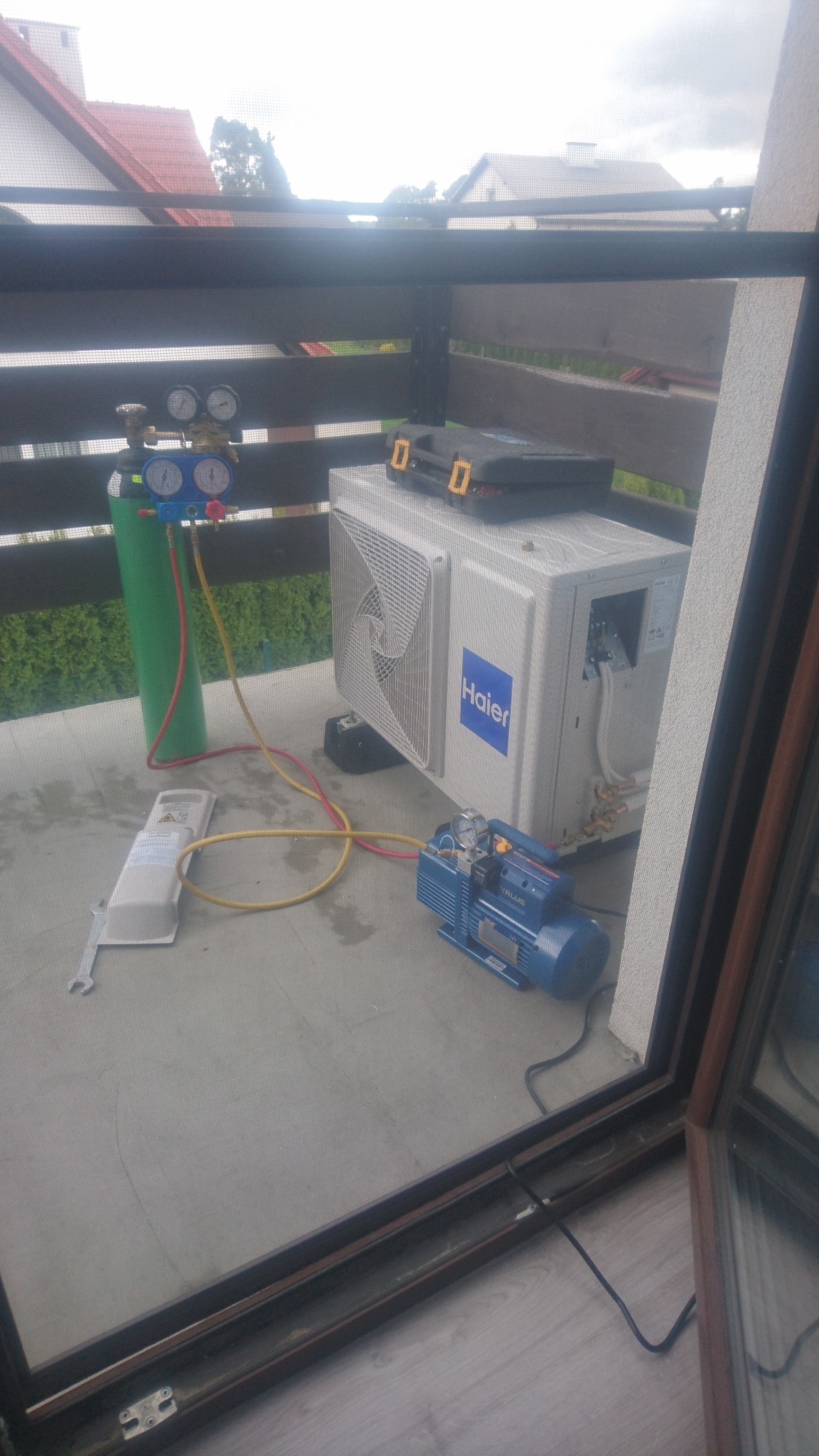 Montaż klimatyzacji Haier na balkonie: widoczny agregat zewnętrzny, butla z czynnikiem chłodniczym, manometry i pompa próżniowa, narzędzia w skrzynce obok.