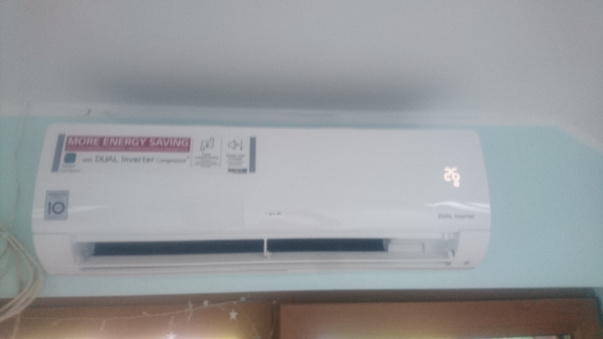 Biały klimatyzator LG Dual Inverter zamontowany na ścianie, wyświetlający temperaturę 26 stopni Celsjusza, z widoczną naklejką 'More Energy Saving' i przewodem zasilającym po lewej stronie.