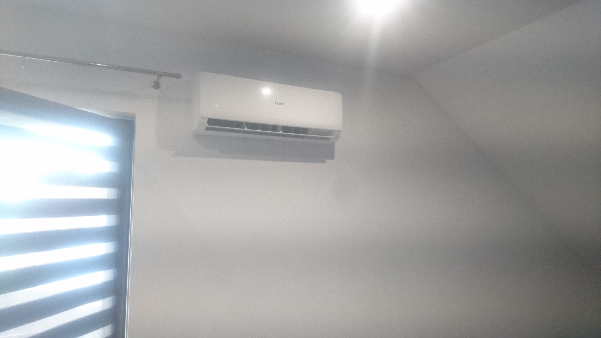 Biała jednostka wewnętrzna klimatyzatora marki Haier zamontowana na jasnej ścianie w pomieszczeniu z roletami okiennymi.