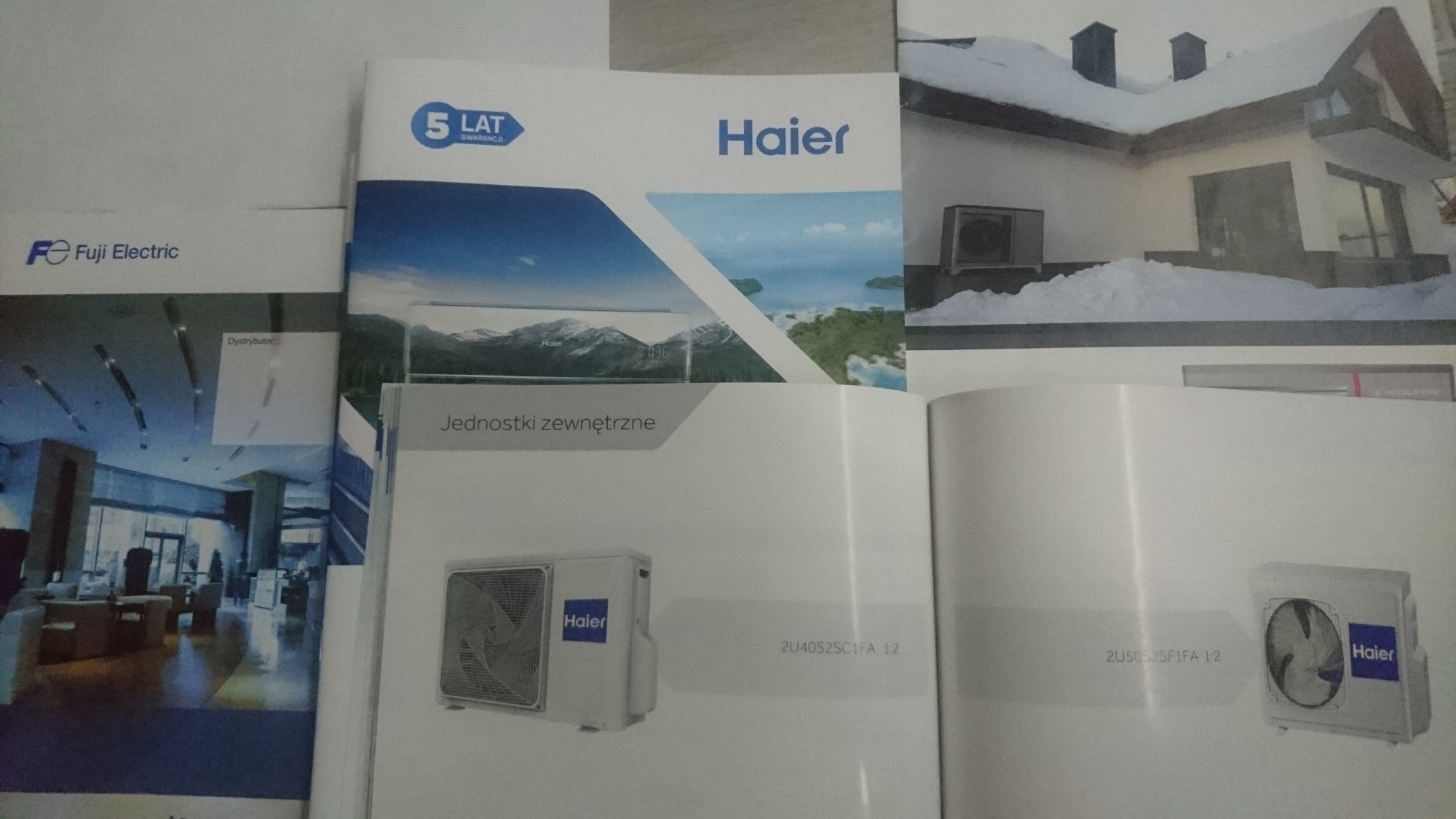Katalogi i ulotki reklamowe różnych producentów urządzeń chłodniczych i wentylacyjnych, w tym Haier i Fuji Electric, prezentujące jednostki zewnętrzne i wnętrza budynków.