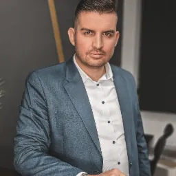 Portret mężczyzny w błękitnej marynarce i białej koszuli, opierającego się o blat. Na nadgarstku widoczny zegarek. Stonowana kolorystyka i profesjonalny wygląd.