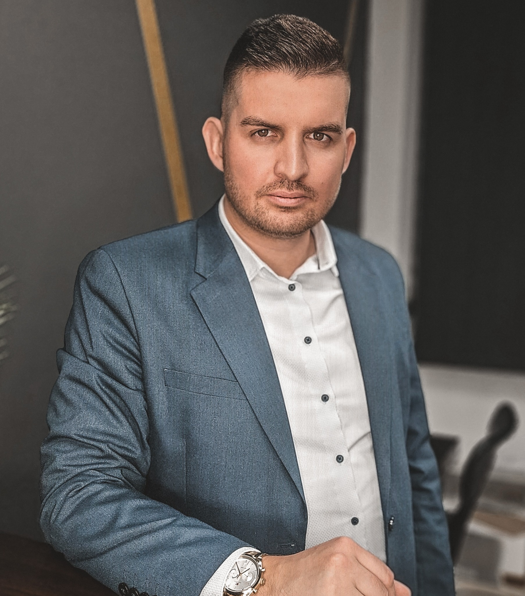 Portret mężczyzny w błękitnej marynarce i białej koszuli, opierającego się o blat. Na nadgarstku widoczny zegarek. Stonowana kolorystyka i profesjonalny wygląd.