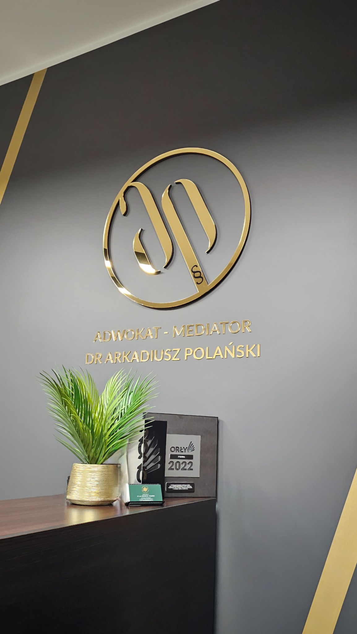 Recepcja kancelarii: złote logo 'AP' na szarym tle, napis 'Adwokat-Mediator Dr Arkadiusz Polański', roślina w doniczce i nagroda 'Orły Prawa 2022'.