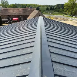 Nowy, szary dach z blachy trapezowej z wyraźnie zaznaczonym kalenicą na tle wiejskich zabudowań i zieleni.