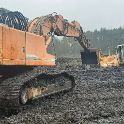 Ciężki sprzęt budowlany, w tym koparka Liebherr 914 i spycharka Liebherr 724, pracują w błotnistym terenie w Podbrzeziu dolnym, prawdopodobnie podczas przygotowywania terenu pod budowę.