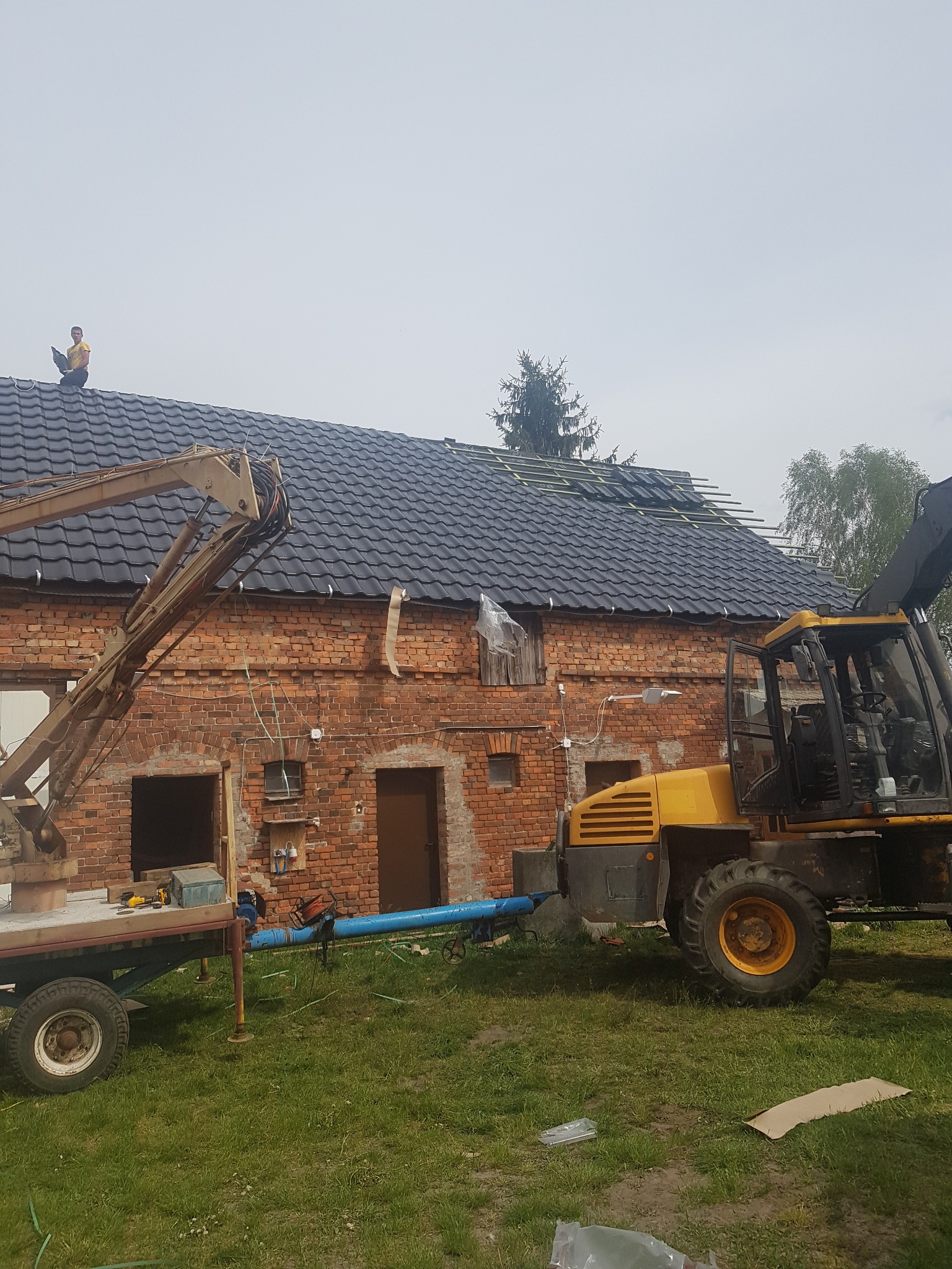 Ceglany budynek z częściowo pokrytym dachem z czarnej dachówki; na dachu pracuje dekarz, a obok stoi żółta maszyna budowlana z podnośnikiem.