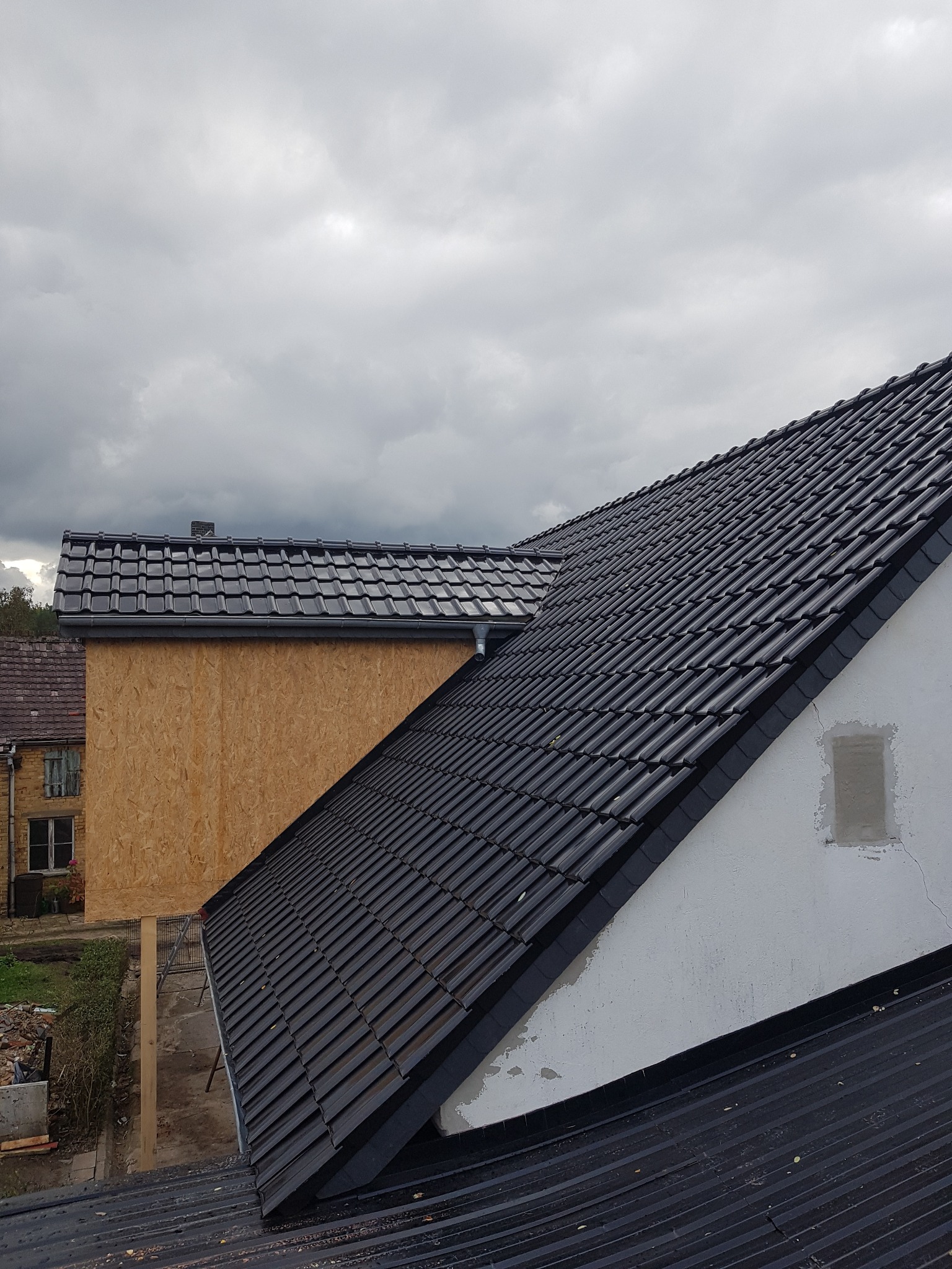 Nowe, błyszczące dachówki ceramiczne na skośnym dachu domu jednorodzinnego, widoczne z góry na tle zachmurzonego nieba i sąsiednich budynków z drewnianą elewacją.