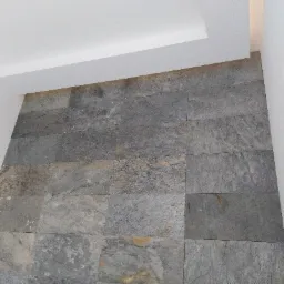 Ściana wykończona szarą, kamienną okładziną w prostokątnych płytkach, z białym sufitem podwieszanym, widok od dołu.