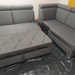 Szara narożna kanapa z funkcją spania, rozłożona na folii ochronnej, widoczne elementy z nadrukiem marki 'Wajnert'.