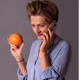 Kobieta z krótkimi włosami w błękitnej koszuli, trzymająca pomarańczę w jednej dłoni i telefon komórkowy w drugiej, zamyślona mina.