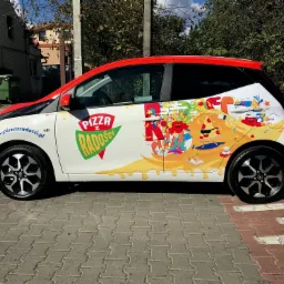 Oklejony folią firmową samochód dostawczy pizzerii 'Pizza z Radości' w Warszawie, z widocznym logo i kolorową grafiką z motywem pizzy i postaci. Na karoserii adres strony internetowej pizzerii.