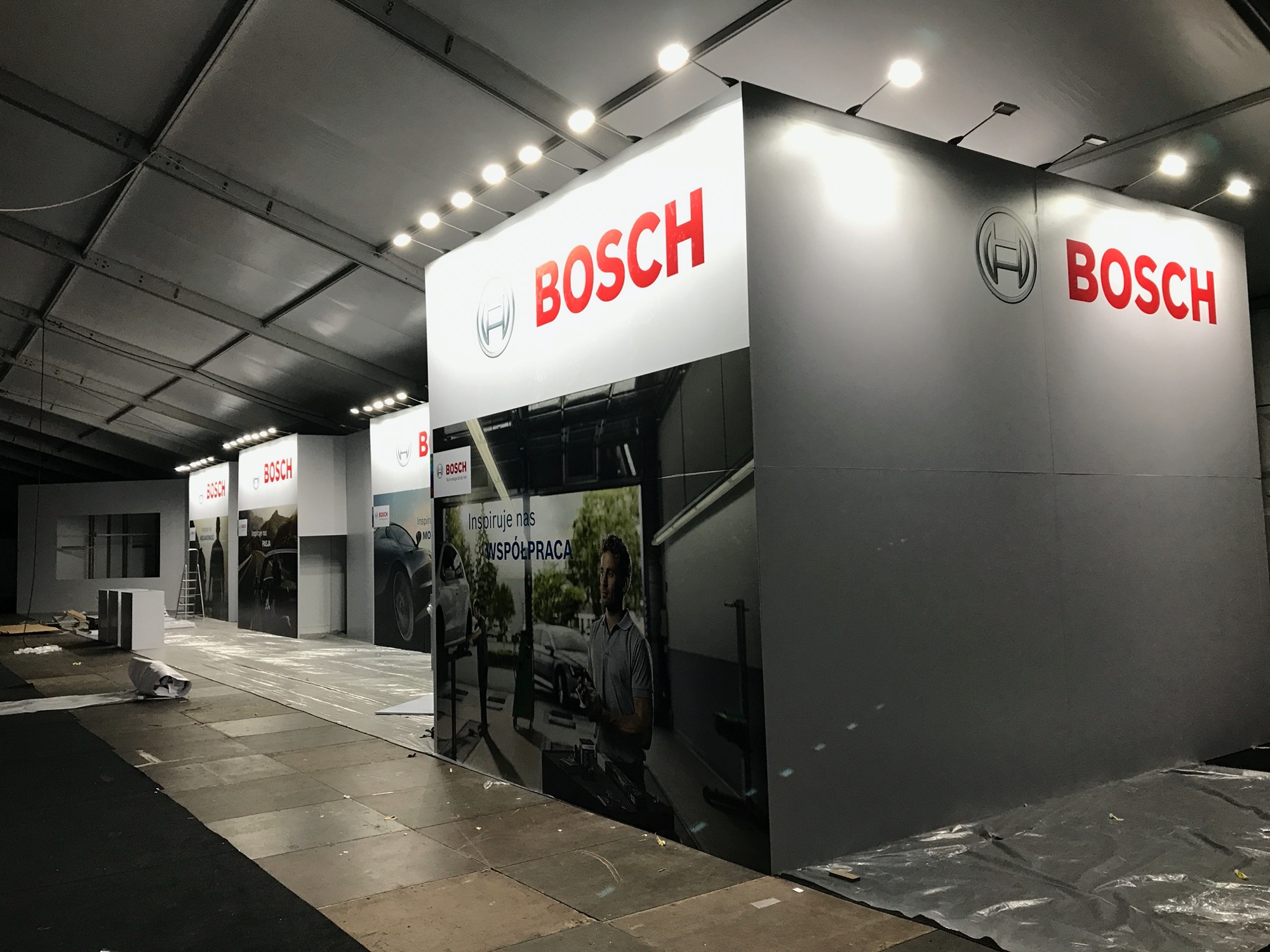 Stoisko targowe z logo Bosch, prezentujące narzędzia i usługi motoryzacyjne, w trakcie przygotowań na tle surowej hali.
