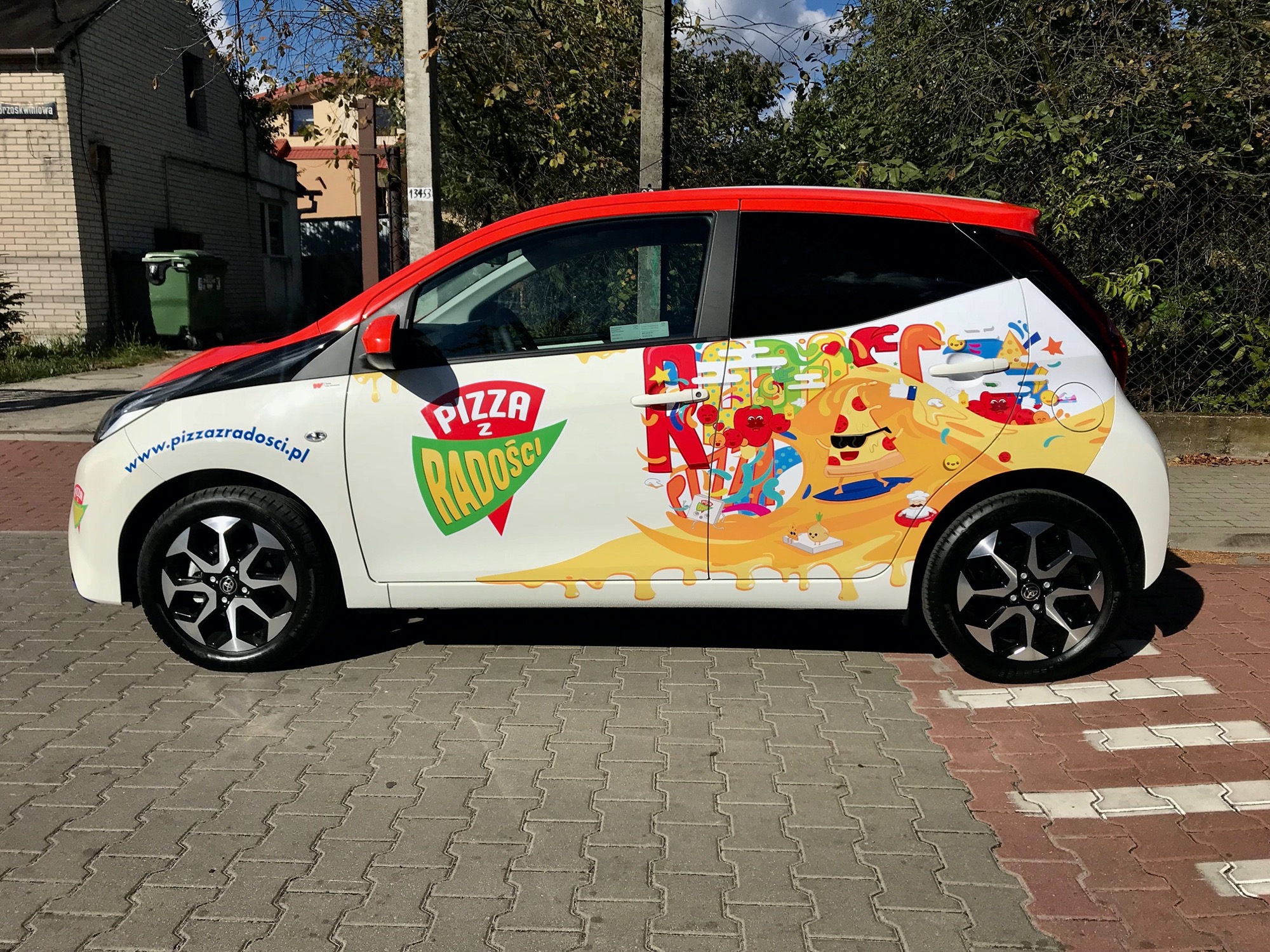Oklejony folią firmową samochód dostawczy pizzerii 'Pizza z Radości' w Warszawie, z widocznym logo i kolorową grafiką z motywem pizzy i postaci. Na karoserii adres strony internetowej pizzerii.