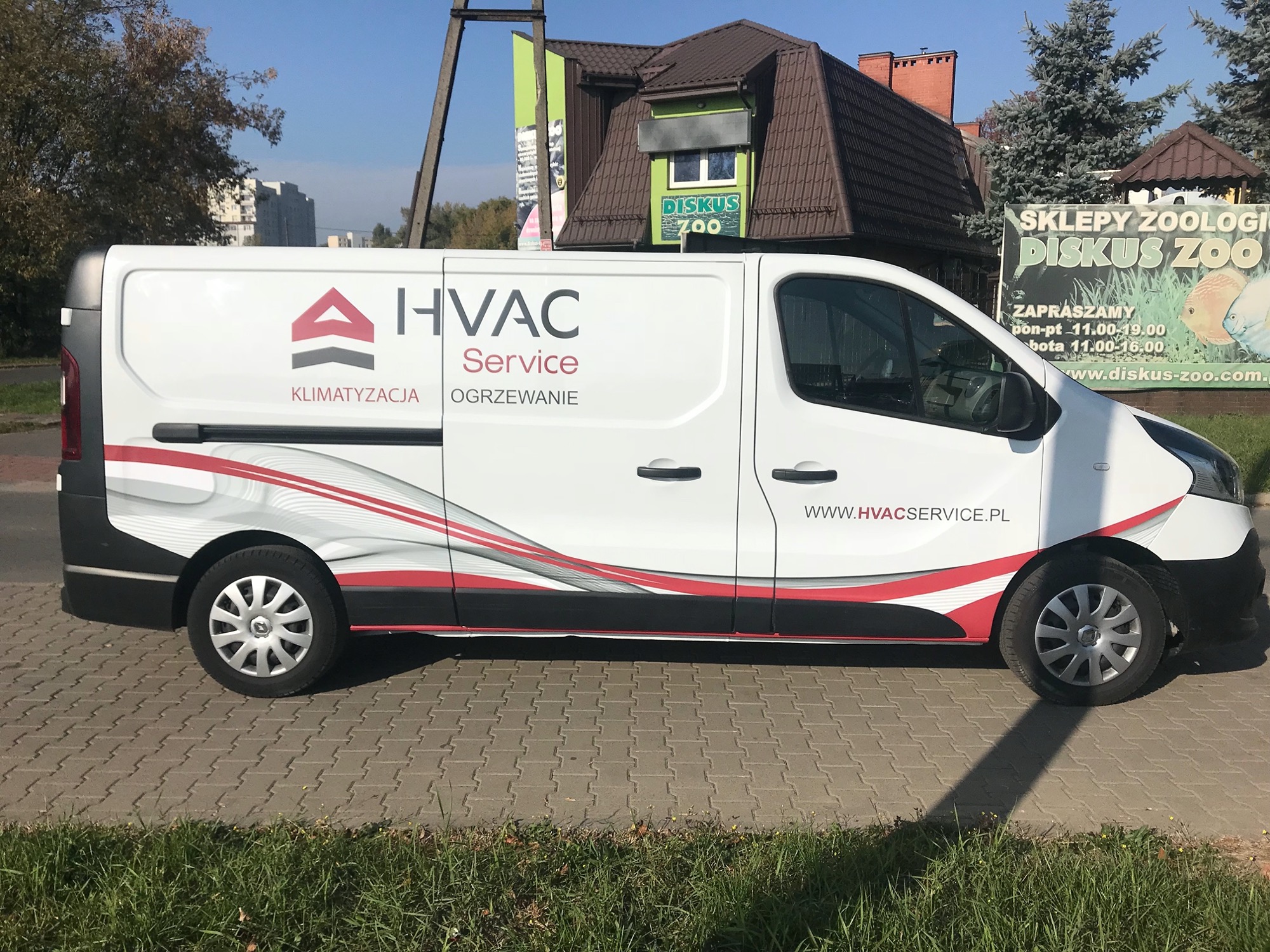 Biały samochód dostawczy z logo firmy HVAC Service (klimatyzacja i ogrzewanie) zaparkowany na chodniku, w tle budynek i szyld sklepu zoologicznego Diskus Zoo.