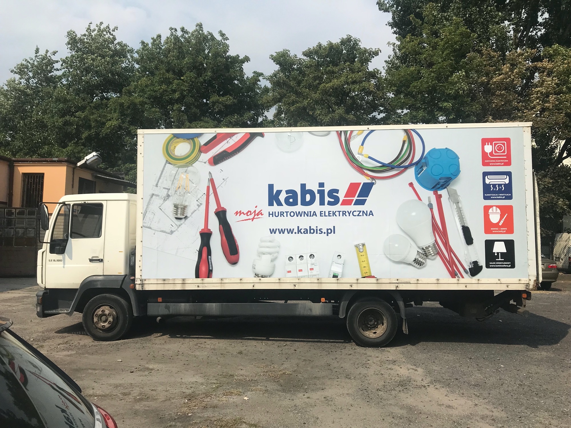 Ciężarówka dostawcza z reklamą hurtowni elektrycznej Kabis, prezentująca narzędzia i materiały elektryczne, takie jak wkrętaki, żarówki LED, miernik, przewody, puszki instalacyjne i schematy...