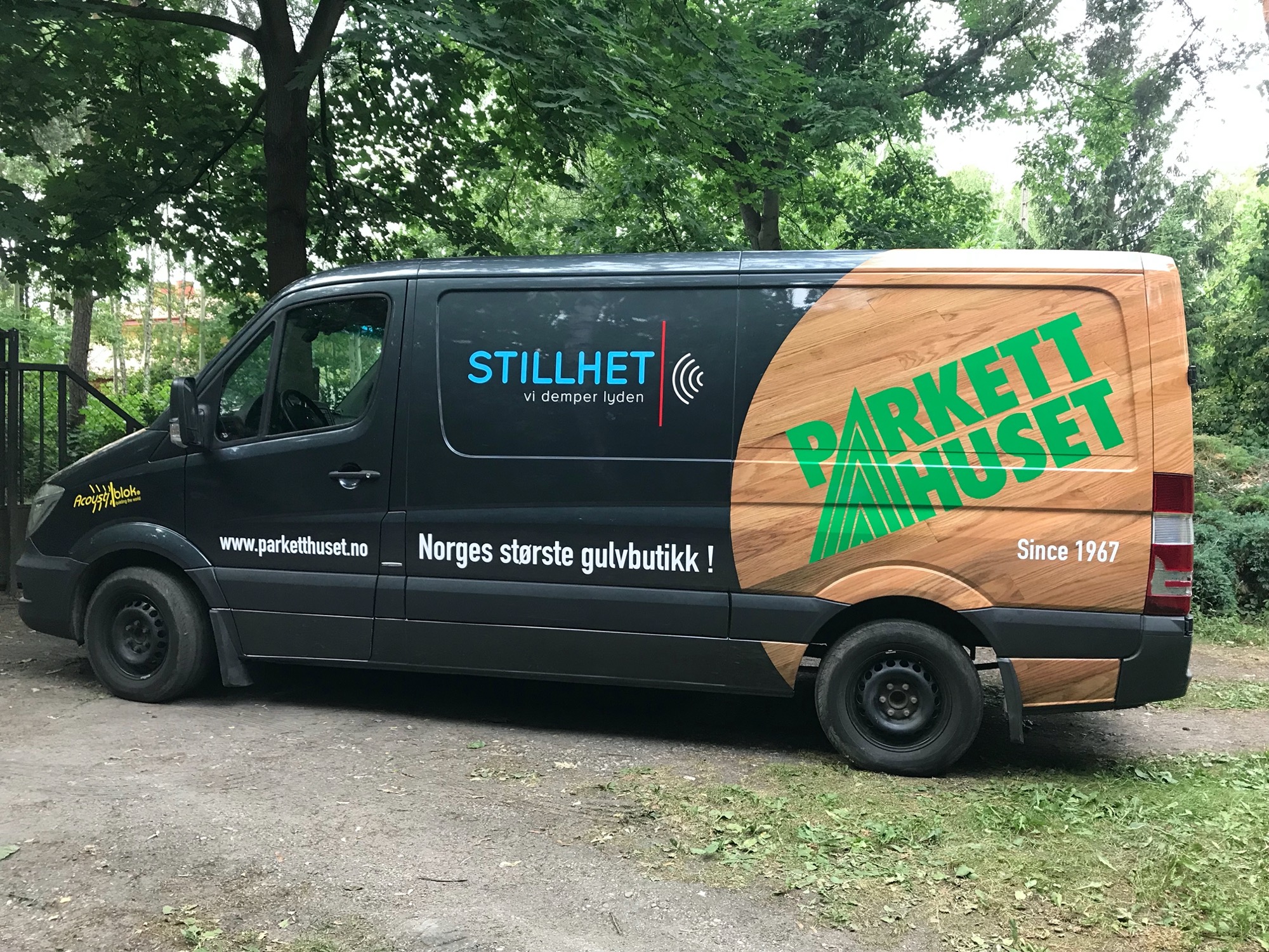 Czarny van z reklamą firmy parkieciarskiej 'Parkett Huset' z datą założenia 1967, zaparkowany na szutrowej drodze wśród zieleni.