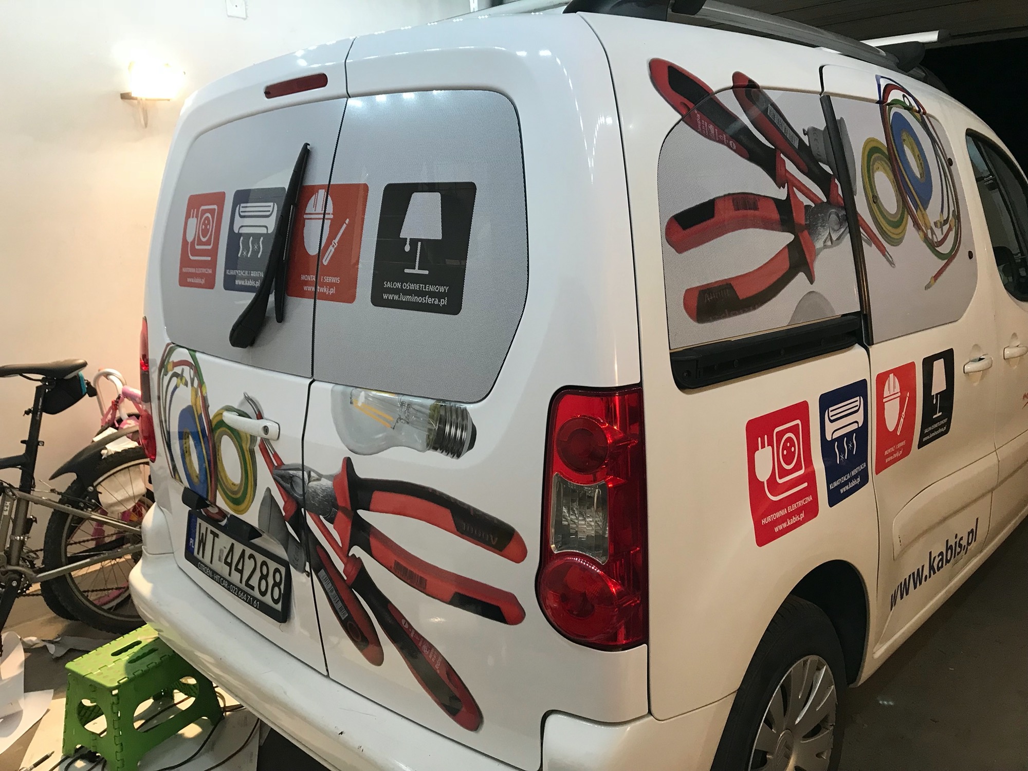 Biały samochód dostawczy z grafiką przedstawiającą narzędzia elektryczne, przewody, żarówkę i logotypy firm z branży elektrycznej, klimatyzacji i oświetlenia.