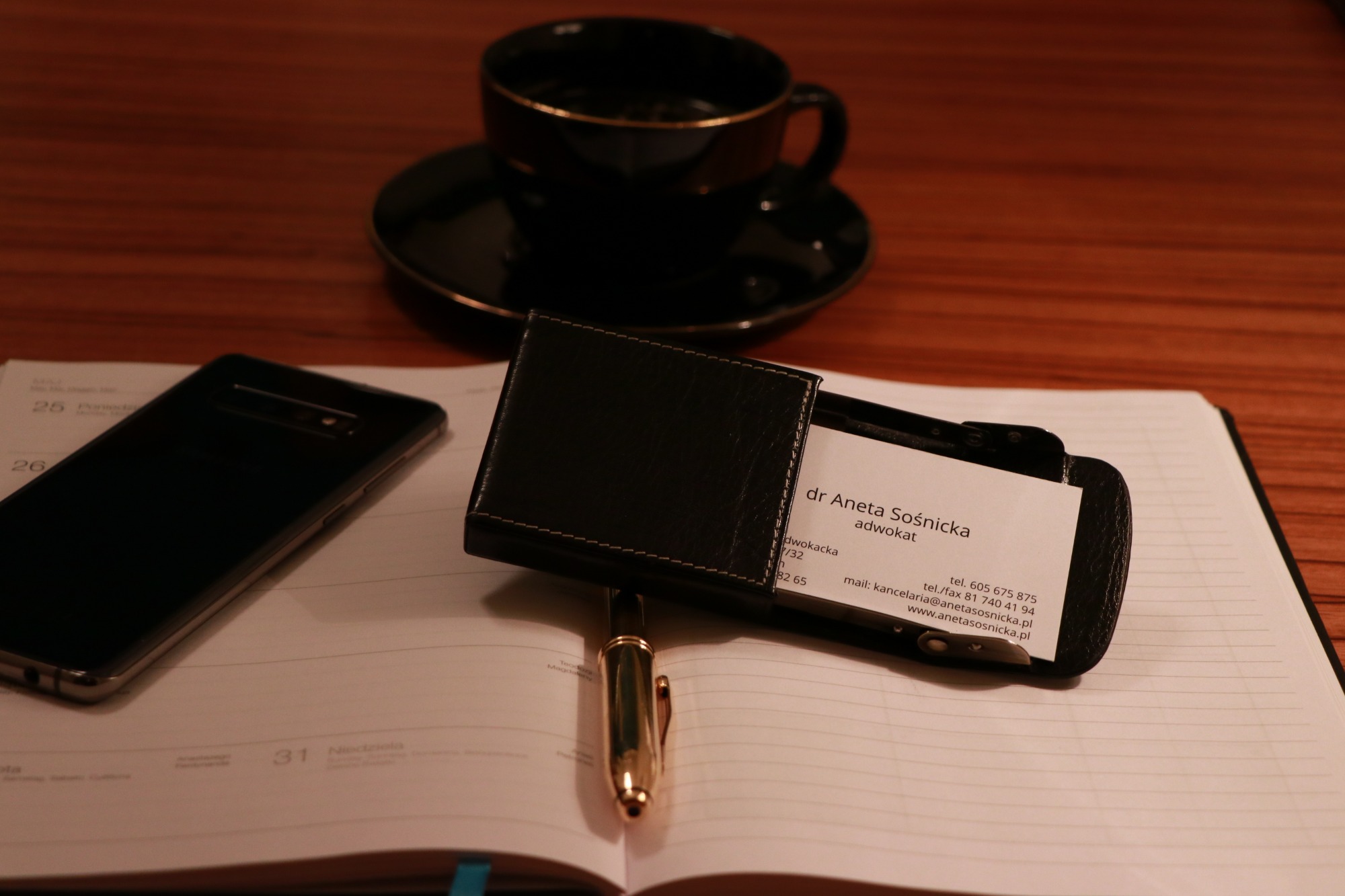 Biurko z kalendarzem, telefonem, piórem i wizytówką adwokat Anety Sośnickiej, na tle filiżanka kawy.
