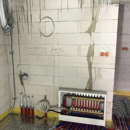 Spectherm sp z o.o. - Rozdzielacz ogrzewania podłogowego w obudowie, rury czerwone i niebieskie ułożone na siatce, białe ściany z bloczków z widocznymi notatkami, manometr na ścianie.