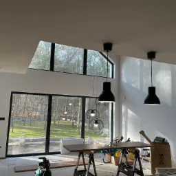 Wnętrze w trakcie wykańczania z dużymi oknami z widokiem na zewnątrz, wiszące czarne lampy nad stołem roboczym, narzędzia i materiały budowlane porozrzucane po podłodze.