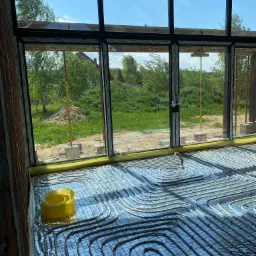 Układanie rur ogrzewania podłogowego na folii aluminiowej w nowym domu z dużymi oknami, widok na zewnątrz na trawnik i drzewa w słoneczny dzień.