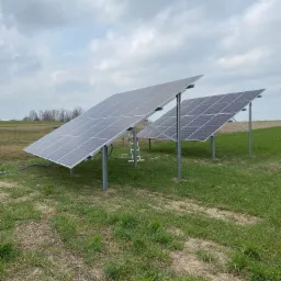 Instalacja paneli fotowoltaicznych na gruncie z widoczną drabiną opartą o konstrukcję, na tle pochmurnego nieba i uprawnego pola.