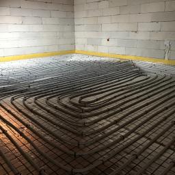 Spectherm sp z o.o. - Układ rur ogrzewania podłogowego rozłożony na folii izolacyjnej z siatką, widoczne ściany z bloczków betonowych i żółta taśma brzegowa.