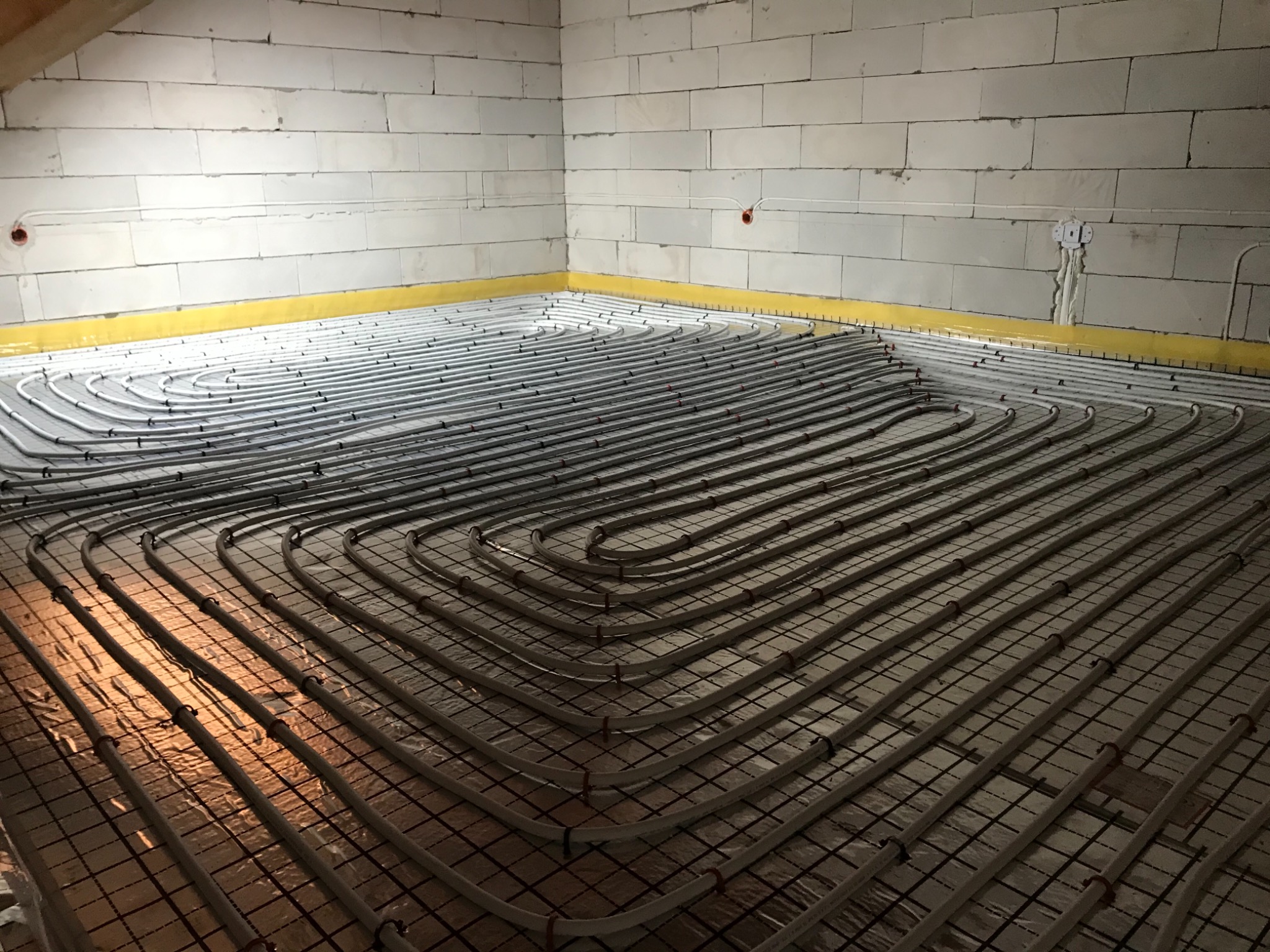 Układ rur ogrzewania podłogowego rozłożony na folii izolacyjnej z siatką, widoczne ściany z bloczków betonowych i żółta taśma brzegowa.