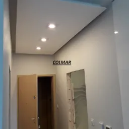 Nowoczesny korytarz z podwieszonym sufitem z płyt kartonowo-gipsowych i wbudowanym oświetleniem punktowym LED, widoczne drzwi wejściowe i przejście do innego pomieszczenia, minimalistyczny design.