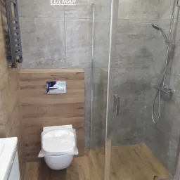 Nowoczesna łazienka z kabiną prysznicową z przezroczystego szkła, podwieszaną miską WC z drewnianą obudową i grzejnikiem drabinkowym w kolorze grafitowym, wykończona płytkami imitującymi kamień...