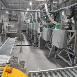 Widok na linię produkcyjną z systemem automatyki przemysłowej, obejmującą przenośniki rolkowe, metalowe szafy sterownicze z zielonymi diodami LED, oraz system rur i przewodów w hali oświetlonej...