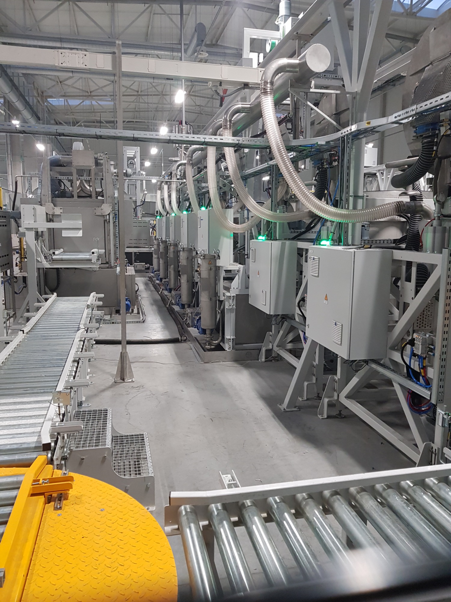 Widok na linię produkcyjną z systemem automatyki przemysłowej, obejmującą przenośniki rolkowe, metalowe szafy sterownicze z zielonymi diodami LED, oraz system rur i przewodów w hali oświetlonej...