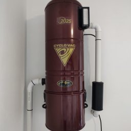 GIT Projekt Paweł Kaczmarzyk - Zainstalowany na białej ścianie, brązowy odkurzacz centralny CycloVac H2025 z widocznymi rurami instalacyjnymi i filtrem.