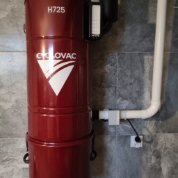 GIT Projekt Paweł Kaczmarzyk - Czerwony odkurzacz centralny CycloVac H725 zamontowany na szarej, marmurkowej ścianie, z widoczną instalacją rurową i gniazdkiem elektrycznym.