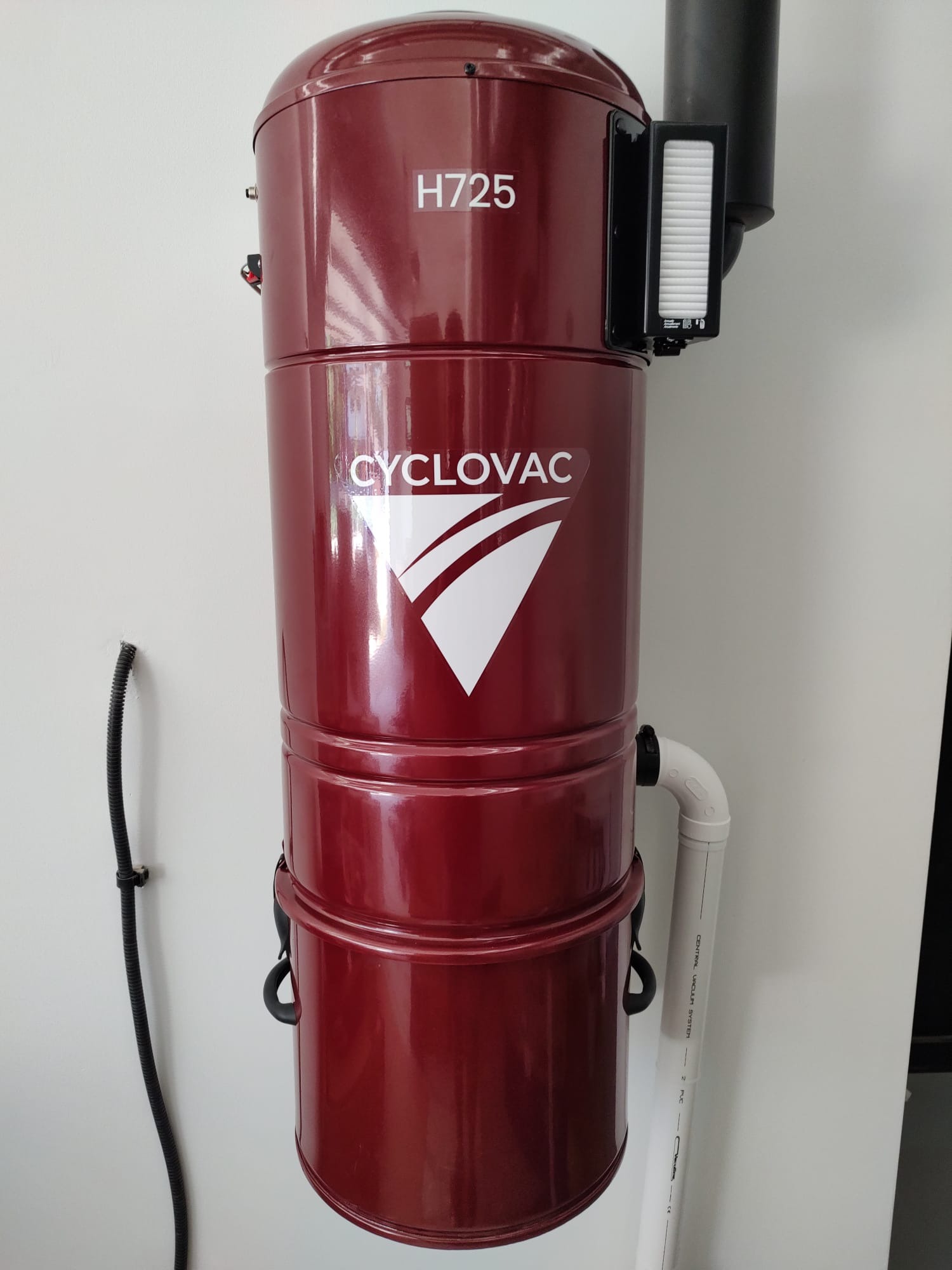 Cylindryczny, bordowy odkurzacz centralny CycloVac H725 zamontowany na białej ścianie, widoczny filtr i rura odprowadzająca.