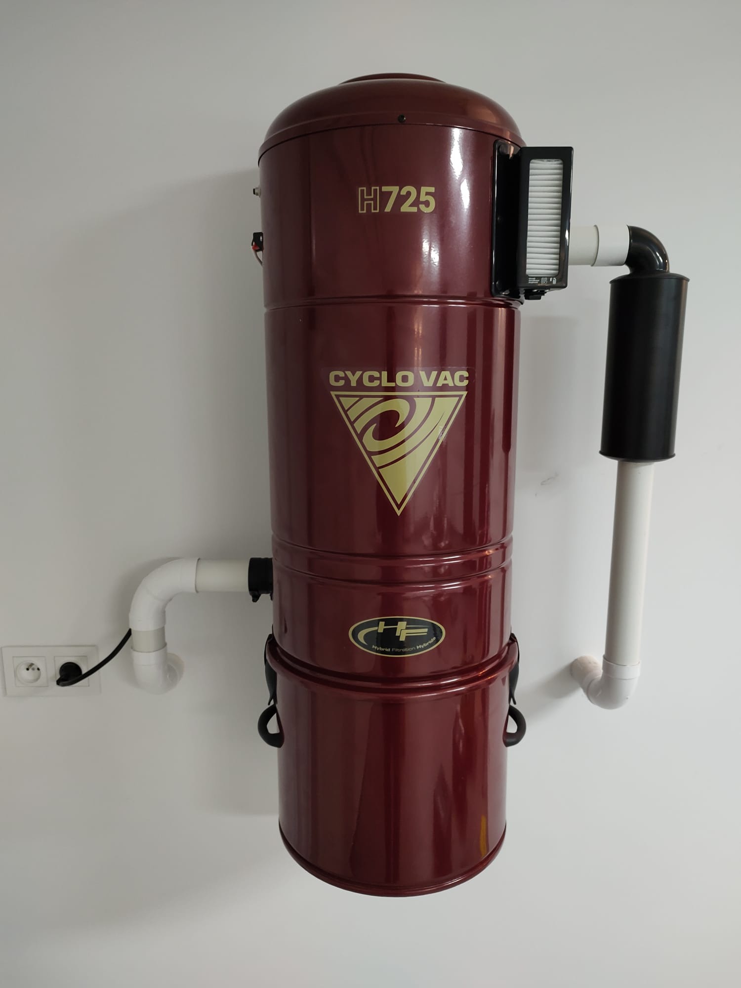 Zamontowany na ścianie, bordowy odkurzacz centralny CycloVac H725 z filtrem i rurami PCV.