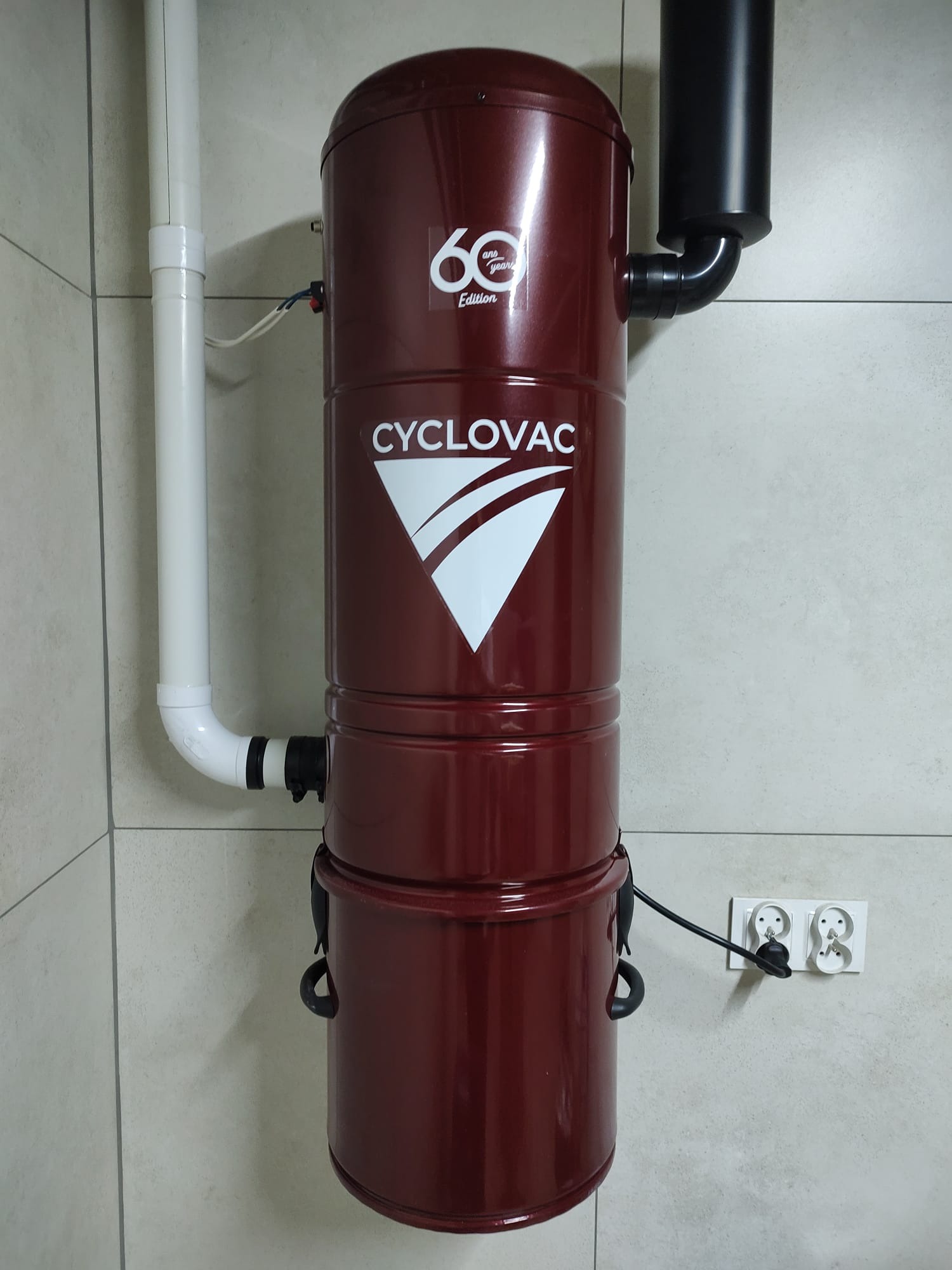 Ciemnoczerwony odkurzacz centralny CycloVac H615 zamontowany na ścianie z widocznymi rurami instalacyjnymi i gniazdkiem elektrycznym.