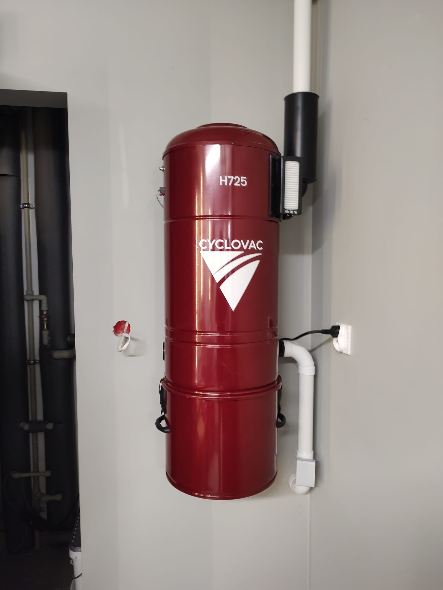 Czerwony, dwuczęściowy model CycloVac H725 zamontowany na szarej ścianie, z widocznymi przewodami i rurami instalacji.