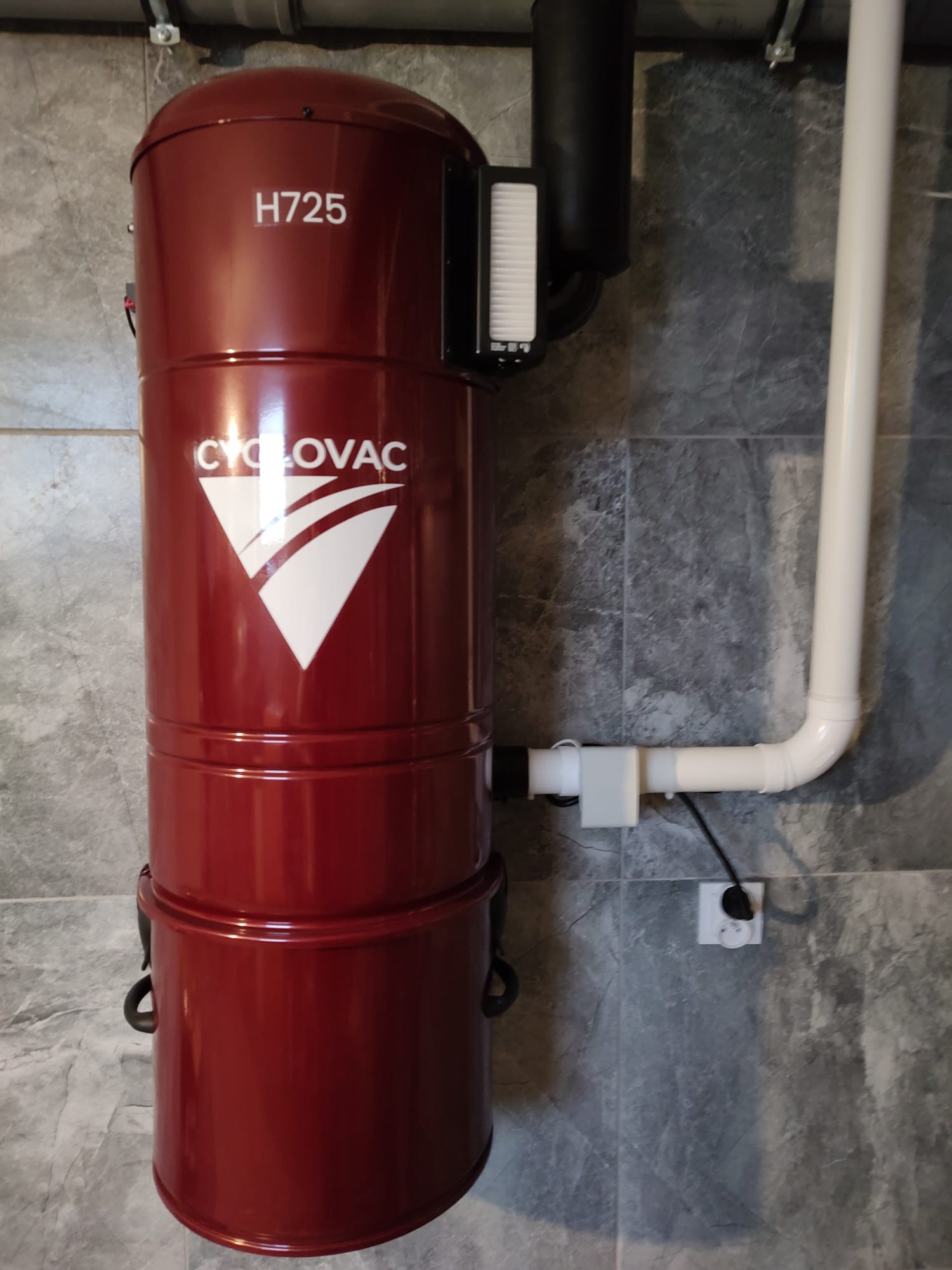 Czerwony odkurzacz centralny CycloVac H725 zamontowany na szarej, marmurkowej ścianie, z widoczną instalacją rurową i gniazdkiem elektrycznym.