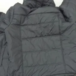 Ciemna, pikowana kurtka z kapturem, widoczna z tyłu, leżąca na jasnym tle. Widoczne regularne przeszycia tworzące poziome pasy. Materiał lekko błyszczący.