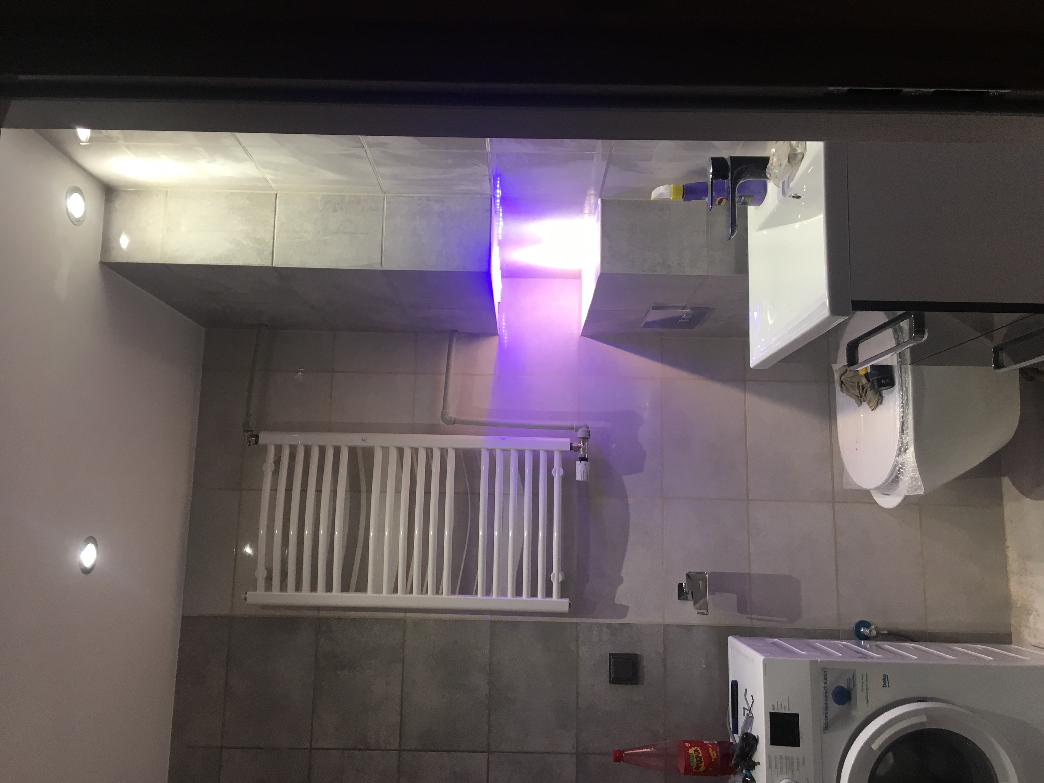 Wykończona łazienka z szarymi płytkami, podświetlanym sufitem, białym grzejnikiem drabinkowym, pralką i umywalką z toaletą w folii ochronnej.