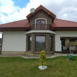 Nowoczesny dom jednorodzinny z jasną elewacją, kamiennym wykończeniem, balkonem i czerwoną dachówką, widok z ogrodu z małym drzewkiem ozdobnym i wózkiem na wąż ogrodowy.