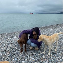 Agnieszka Jednoróg - Kobieta w fioletowej kurtce kuca na kamienistej plaży, obejmując brązowego labradora, obok stoi beżowy labrador, w tle pochmurne niebo i morze z widocznym statkiem.