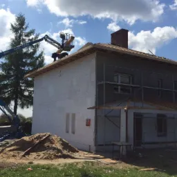 Dwukondygnacyjny dom w trakcie budowy z częściowo otynkowaną elewacją, rusztowaniami i podnośnikiem koszowym, na którym pracują dwie osoby przy przygotowaniu dachu, widoczny stos piasku.